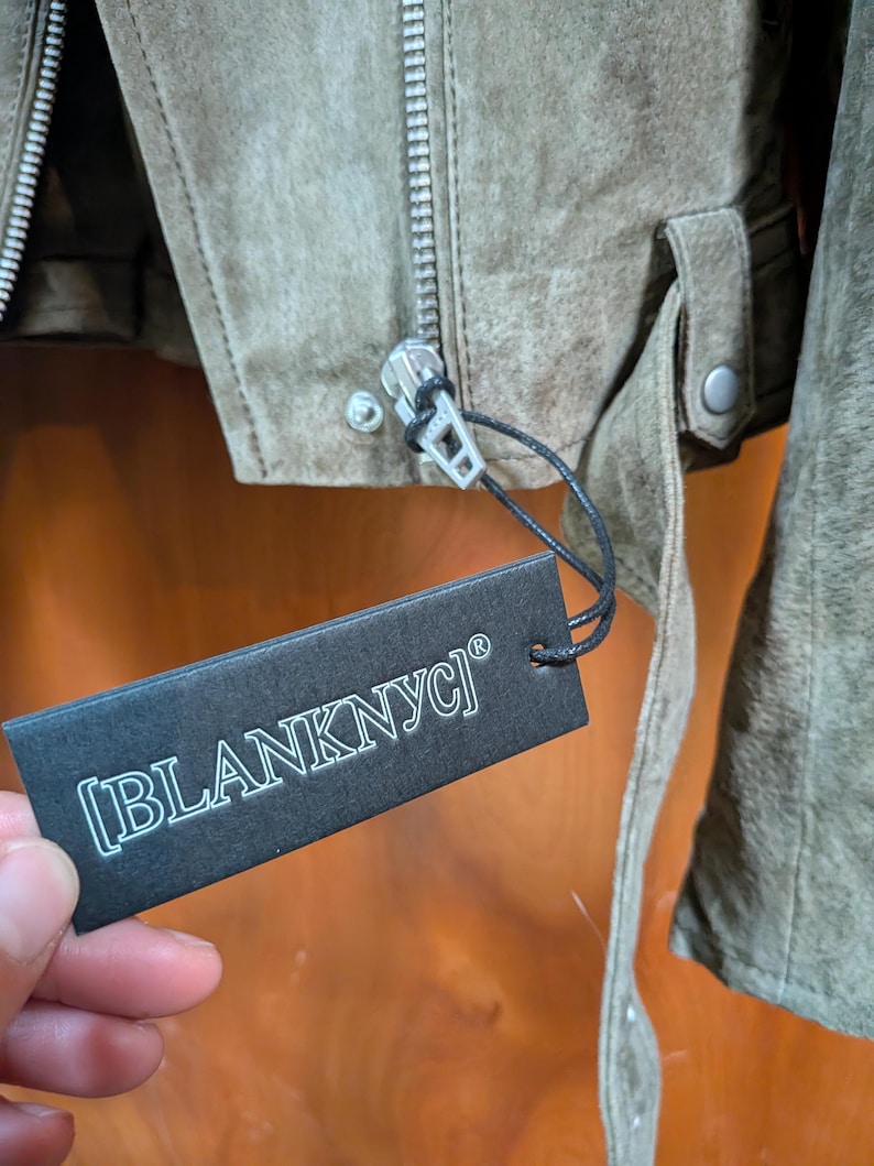 May include: Close-up of a suede jacket in a muted olive green color. The jacket features a silver zipper, a small strap with a button, and a tag that reads "[BLANKNYC]" in white text.