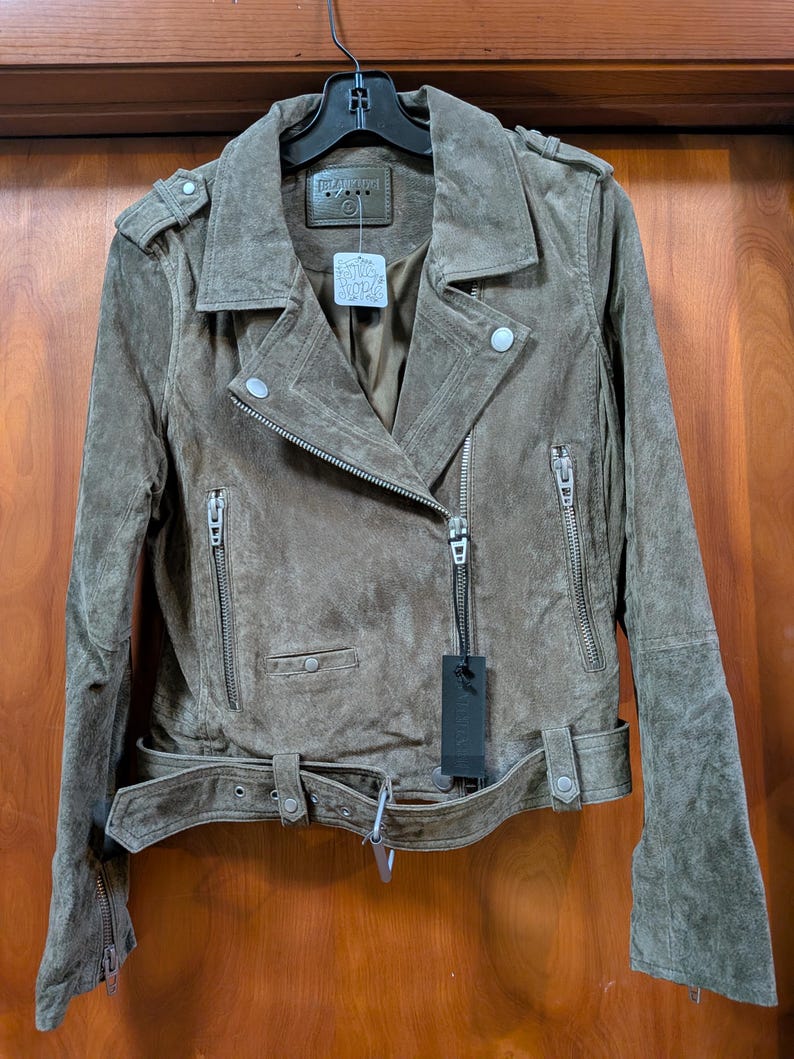May include: A brown suede jacket with a classic biker style. Features include an asymmetrical zipper closure, multiple zippered pockets, a belted waist, and shoulder epaulets. The jacket is hanging on a black hanger against a wooden door.