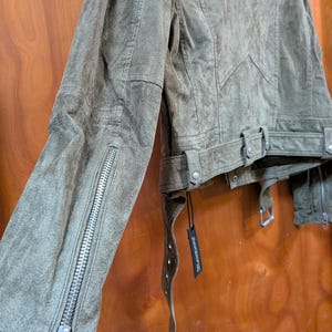 May include: Olive green suede jacket with silver zipper details on the sleeves. The jacket features a belted waist and a classic biker jacket design. The suede has a distressed look, and a black tag hangs from the jacket.
