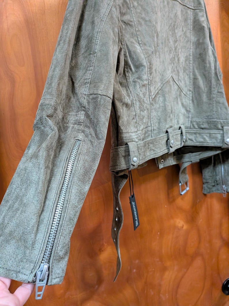 May include: Olive green suede jacket with silver zipper details on the sleeves. The jacket features a belted waist and a classic biker jacket design. The suede has a distressed look, and a black tag hangs from the jacket.