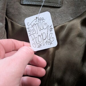 May include: Olive green jacket with a satin lining. The tag reads "Free People" in a handwritten style. The jacket has a leather label that reads "Blank NYC".