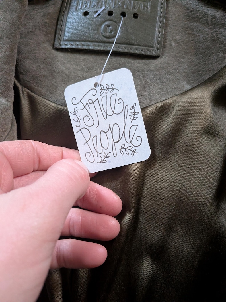 May include: Olive green jacket with a satin lining. The tag reads "Free People" in a handwritten style. The jacket has a leather label that reads "Blank NYC".