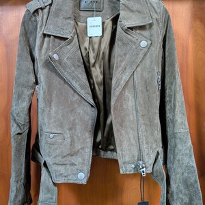 May include: A brown suede jacket with a classic biker style. Features include an asymmetrical zipper, notched lapels, and a belt at the waist. The jacket has silver hardware and a soft, textured appearance. The jacket is hanging on a black hanger.