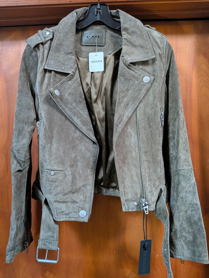 May include: A brown suede jacket with a classic biker style. Features include an asymmetrical zipper, notched lapels, and a belt at the waist. The jacket has silver hardware and a soft, textured appearance. The jacket is hanging on a black hanger.