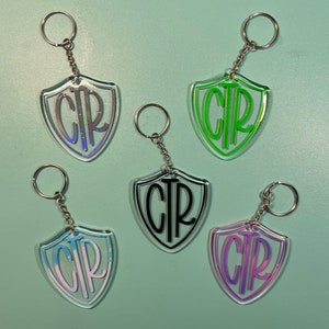 May include: Five clear acrylic keychain charms shaped like shields with the initials "CTR" in different colors. The initials are in black, green, pink, purple, and a rainbow iridescent color.
