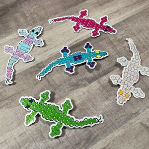 Pode incluir: Cinco adesivos de lagarto de contas coloridas. Os lagartos s&atilde;o feitos de pequenas contas de v&aacute;rias cores, incluindo rosa, azul, verde, branco e vermelho. Os adesivos s&atilde;o perfeitos para adicionar um toque caprichoso a qualquer superf&iacute;cie.