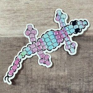 Pode incluir: Um lagarto colorido feito de contas Perler. O lagarto &eacute; rosa, azul e verde com um contorno preto. As contas s&atilde;o dispostas em um padr&atilde;o para criar a forma de um lagarto.