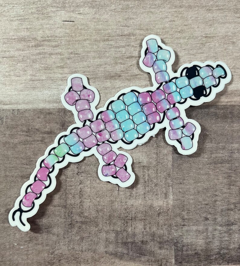 Pode incluir: Um lagarto colorido feito de contas Perler. O lagarto &eacute; rosa, azul e verde com um contorno preto. As contas s&atilde;o dispostas em um padr&atilde;o para criar a forma de um lagarto.