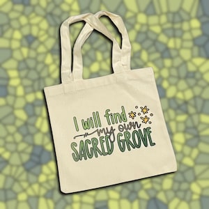 May include: Cream-colored tote bag with the text "I will find my own Sacred Grove" in green and purple. The bag has two cream-colored handles and a simple design, suitable for carrying books or groceries.