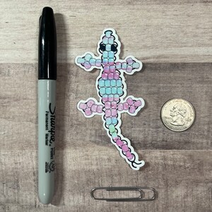 Pode incluir: Um adesivo de um lagarto rosa, azul e verde feito de pequenas contas. O lagarto est&aacute; virado para a direita e tem um olho preto.