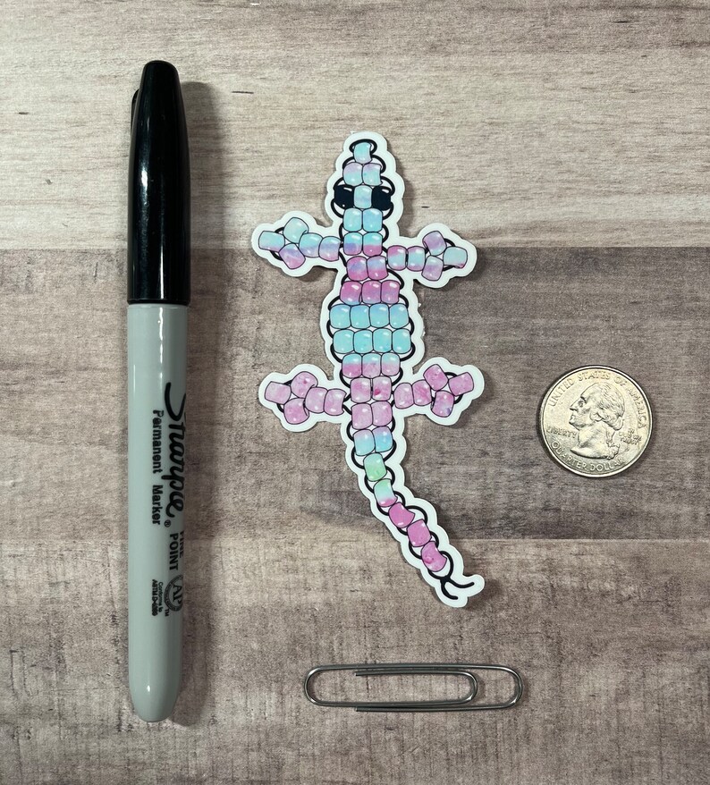 Pode incluir: Um adesivo de um lagarto rosa, azul e verde feito de pequenas contas. O lagarto est&aacute; virado para a direita e tem um olho preto.
