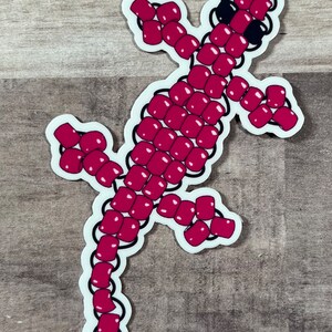 Pode incluir: Um adesivo de lagarto rosa de contas. O lagarto est&aacute; em uma pose de caminhada com o rabo enrolado atr&aacute;s dele.
