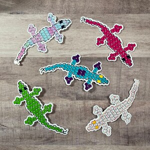 Pode incluir: Conjunto de cinco adesivos de lagarto coloridos feitos de contas de pl&aacute;stico. Os lagartos s&atilde;o de v&aacute;rias cores, incluindo rosa, azul, verde, branco e vermelho.