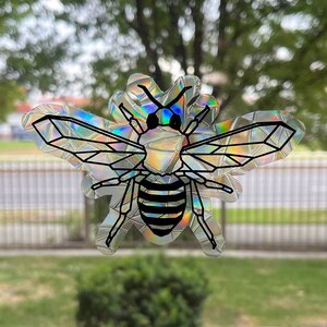 May include: A holographic bee sticker with a black outline and iridescent wings. The bee has a black and white striped body.