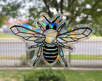 Pode incluir: Um adesivo de abelha holográfico com contorno preto e asas iridescentes. A abelha tem um corpo listrado de preto e branco.