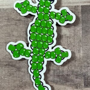 Pode incluir: Adesivo de lagarto de contas verdes. O lagarto &eacute; feito de contas verdes e tem olhos pretos. O adesivo est&aacute; em um fundo de madeira.