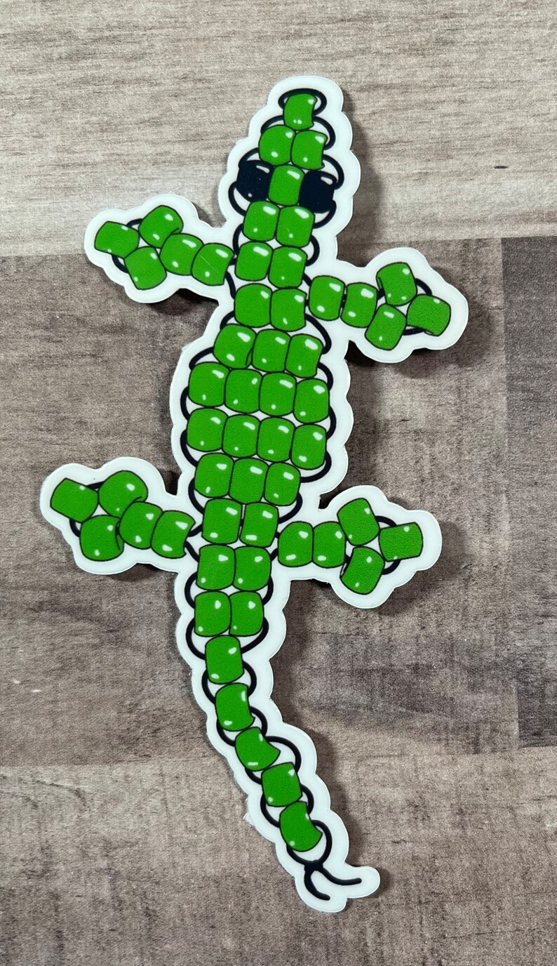 Pode incluir: Adesivo de lagarto de contas verdes. O lagarto &eacute; feito de contas verdes e tem olhos pretos. O adesivo est&aacute; em um fundo de madeira.