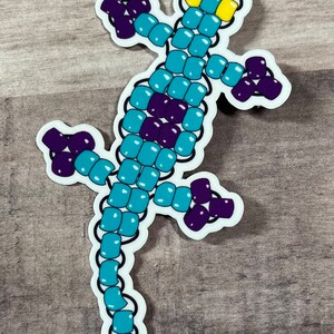 Pode incluir: Um adesivo de lagarto de contas turquesa e roxo. O lagarto est&aacute; em p&eacute; sobre suas patas traseiras com o rabo enrolado atr&aacute;s dele. O lagarto tem olhos amarelos.