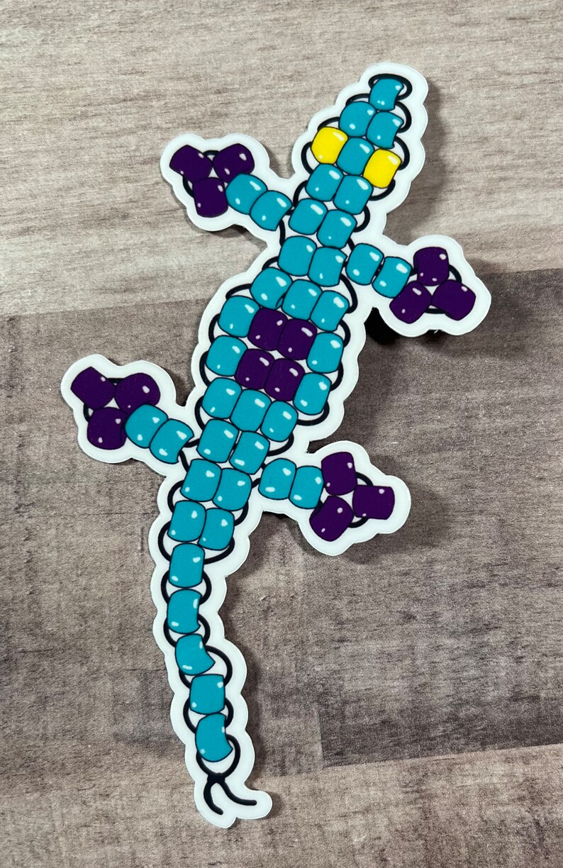 Pode incluir: Um adesivo de lagarto de contas turquesa e roxo. O lagarto est&aacute; em p&eacute; sobre suas patas traseiras com o rabo enrolado atr&aacute;s dele. O lagarto tem olhos amarelos.