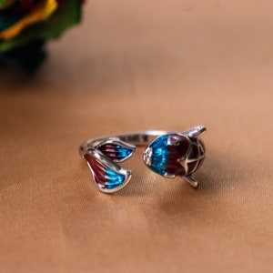 Può includere: Un anello d'argento con un design a forma di pesce in smalto blu e bordeaux. Il pesce ha un design dettagliato e una piccola pietra rossa per l'occhio.