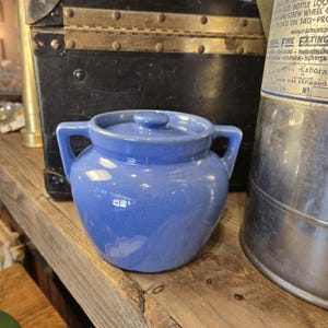May include: A periwinkle blue ceramic sugar bowl with a matching lid and two side handles. The bowl has a rounded, bulbous body and a smooth, glossy finish. The lid features a small, centered knob handle for easy removal from the container.