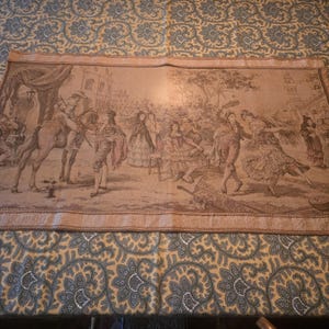 May include: A tapestry depicting a historical scene with figures in period clothing, including a man on horseback and people dancing. The tapestry is primarily beige and brown, with a detailed design and a decorative border. The tapestry is displayed on a patterned fabric.