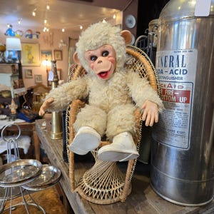 May include: A plush monkey doll with cream-colored fur, a vinyl face, and white shoes, seated in a small wicker peacock chair. Next to the chair is a large, cylindrical metal General Soda-Acid fire extinguisher with a prominent label.