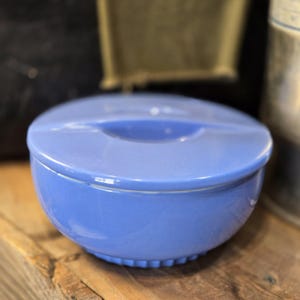 May include: A round, periwinkle blue ceramic bowl with a matching lid. The bowl features a smooth, glossy finish and a subtle, ribbed texture along the bottom edge. The lid has a recessed, circular handle integrated into its center design.