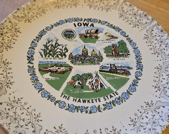 May include: A vintage decorative plate featuring the state of Iowa. The plate has a white background with a gold floral border. The center displays Iowa landmarks and the text "IOWA" and "THE HAWKEYE STATE".