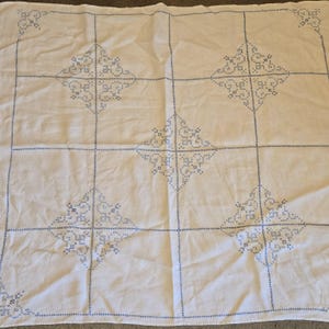 May include: White tablecloth with blue embroidered geometric patterns. The design features a grid of squares, each containing a diamond-shaped motif. The embroidery is detailed and evenly spaced, creating a classic, handmade aesthetic.