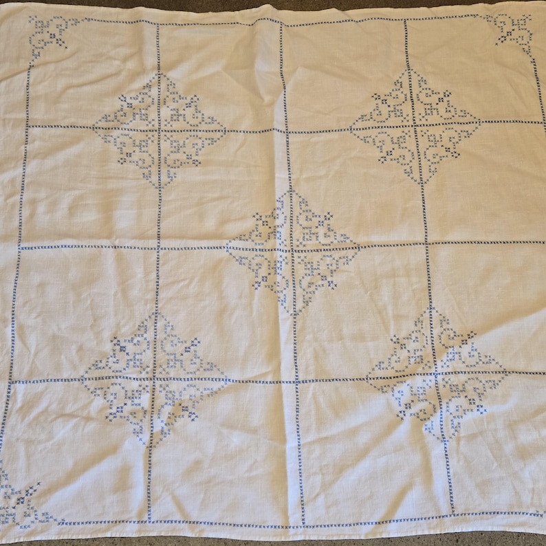 May include: White tablecloth with blue embroidered geometric patterns. The design features a grid of squares, each containing a diamond-shaped motif. The embroidery is detailed and evenly spaced, creating a classic, handmade aesthetic.
