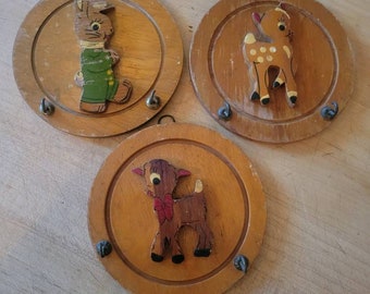 May include: Three wooden wall-mounted coat racks with animal designs. The first rack features a rabbit wearing green overalls, the second rack features a deer with white spots, and the third rack features a deer with a red bow.