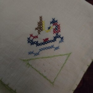 Puede incluir: Toalla de t&eacute; de lino blanco con un tri&aacute;ngulo bordado verde y un dise&ntilde;o de punto de cruz colorido de un p&aacute;jaro en el centro.