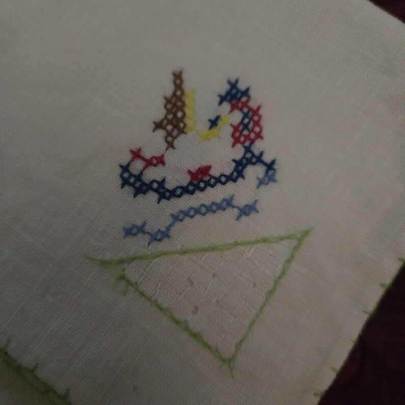 Puede incluir: Toalla de t&eacute; de lino blanco con un tri&aacute;ngulo bordado verde y un dise&ntilde;o de punto de cruz colorido de un p&aacute;jaro en el centro.