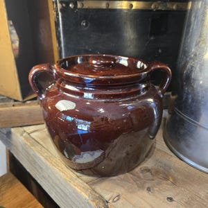 May include: A dark brown glazed ceramic pot with a matching lid and two side handles. The pot features a rounded body and a smooth, reflective surface finish. The lid has a small central knob for lifting, and the overall design is simple.