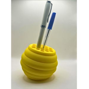 May include: Yellow honeycomb-shaped desk organiser with two pens, one white and one blue, standing upright in the holes.