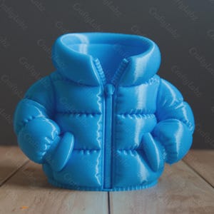 May include: A light blue, 3D-printed miniature puffer jacket. The jacket features a full zipper, a high collar, and two front pockets. The design mimics a real jacket, with quilted sections and puffy sleeves. The object is displayed on a wooden surface.