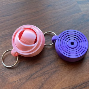 May include: Two keychains, one pink and one purple. The pink keychain is a sphere with a smaller sphere inside. The purple keychain is a flat circle with concentric rings.