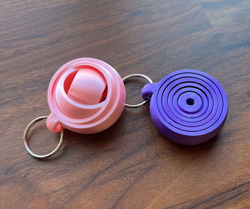 May include: Two keychains, one pink and one purple. The pink keychain is a sphere with a smaller sphere inside. The purple keychain is a flat circle with concentric rings.
