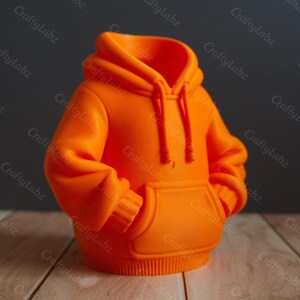 May include: An orange hoodie-shaped container with a hood, drawstrings, and a front pocket. The container is made of a solid material and has a smooth finish. The design is a miniature replica of a hoodie.