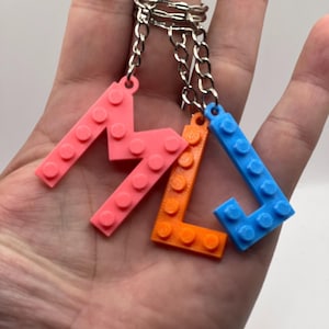 May include: Three plastic letter charms in the shape of the letters M, L, and J. The charms are attached to silver keychains. The M is pink, the L is orange, and the J is blue.
