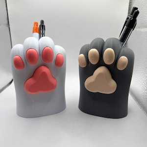 May include: Two cat paw-shaped pen holders. One is white with pink paw pads, and the other is dark grey with tan paw pads. Pens are inserted into the holders. These desk organisers are a fun way to store pens and pencils.