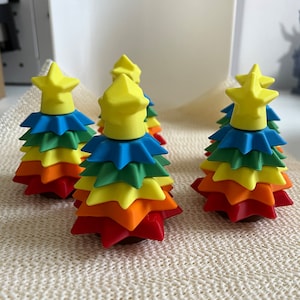 May include: Five rainbow-coloured Christmas tree decorations with a yellow star on top. Each decoration is made of stacked star-shaped pieces in red, orange, yellow, green, blue, and purple.