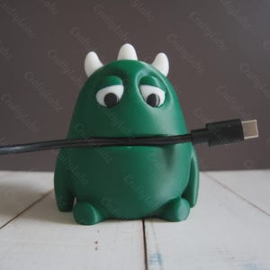 May include: A green monster-shaped cable tidy with white horns and sad eyes, holding a black USB-C cable. The product is on a white wooden surface with a dark grey background. The product is labelled with the text "CraftyLabz".