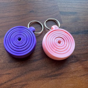 May include: Two keychains with a circular design. One keychain is purple with a silver ring and the other is pink with a silver ring.