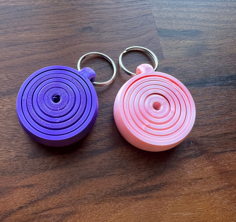 May include: Two keychains with a circular design. One keychain is purple with a silver ring and the other is pink with a silver ring.