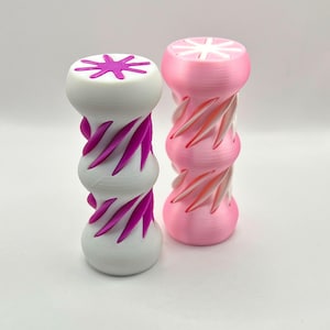 以下が含まれることがあります： 2つの装飾的な円筒形のオブジェクト。1つは白で、マゼンタのアクセントと上部にスターバーストのデザインがあります。もう1つはピンクで、白と赤のアクセントがあります。どちらもねじれたリブのデザインです。