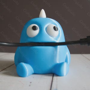 May include: A light blue, cartoon-style cable tidy with large white-rimmed eyes and a small white horn. The tidy has a rounded body and small, rounded feet. A black cable is inserted through the tidy.