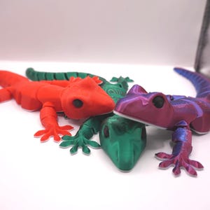 May include: Three articulated lizard figurines in red, green, and a purple-blue gradient. The lizards are posed on a white surface, showcasing their detailed design and segmented bodies. These decorative figurines are likely 3D-printed.