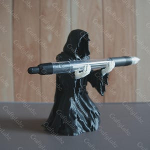 May include: A black Grim Reaper figure holding a large clear pen. The pen has black ends and a clear barrel. The Reaper is wearing a black hooded robe and has white skeletal hands. The CraftyLabz logo is visible.